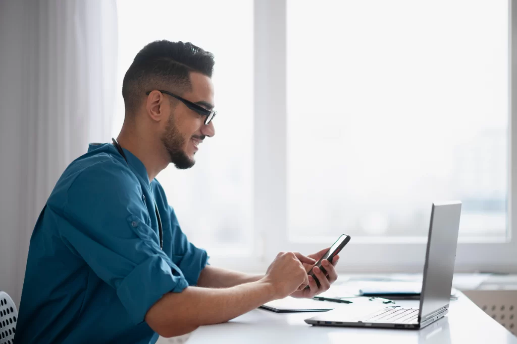 Homem de negócios em frente a um notebook, olhando o celular, ilustrando o tema de obrigações fiscais e tarefas administrativas para pequenas e médias empresas (PMEs).