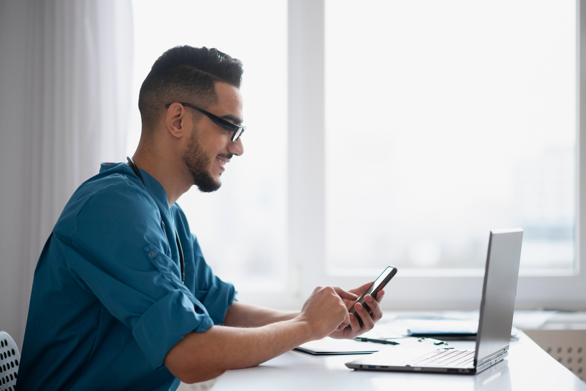 Homem de negócios em frente a um notebook, olhando o celular, ilustrando o tema de obrigações fiscais e tarefas administrativas para pequenas e médias empresas (PMEs).