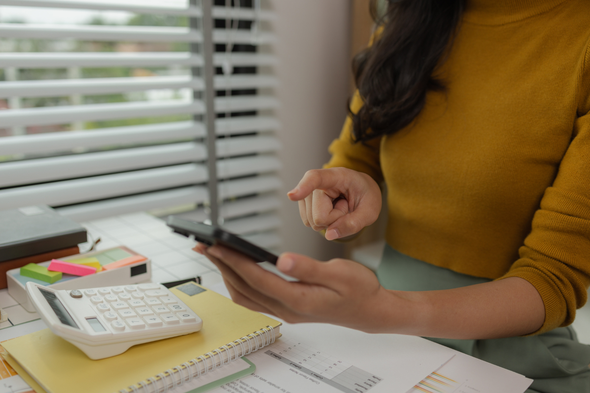Empresária em ambiente de escritório, lendo informações no celular com documentos, calculadora e cadernos sobre a mesa, representando empresa em processo de emissão de nota fiscal por prestação ou subcontratação de serviços.
