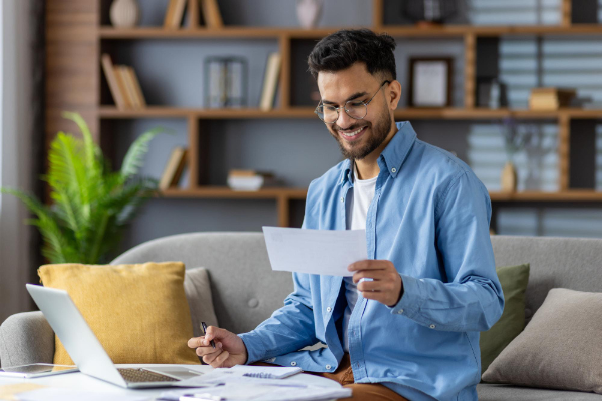 Homem com um documento em mãos sentado em frente o notebook lendo artigo sobre emissão de nota fiscal em contingência: o que é, quando usar e como regularizar.