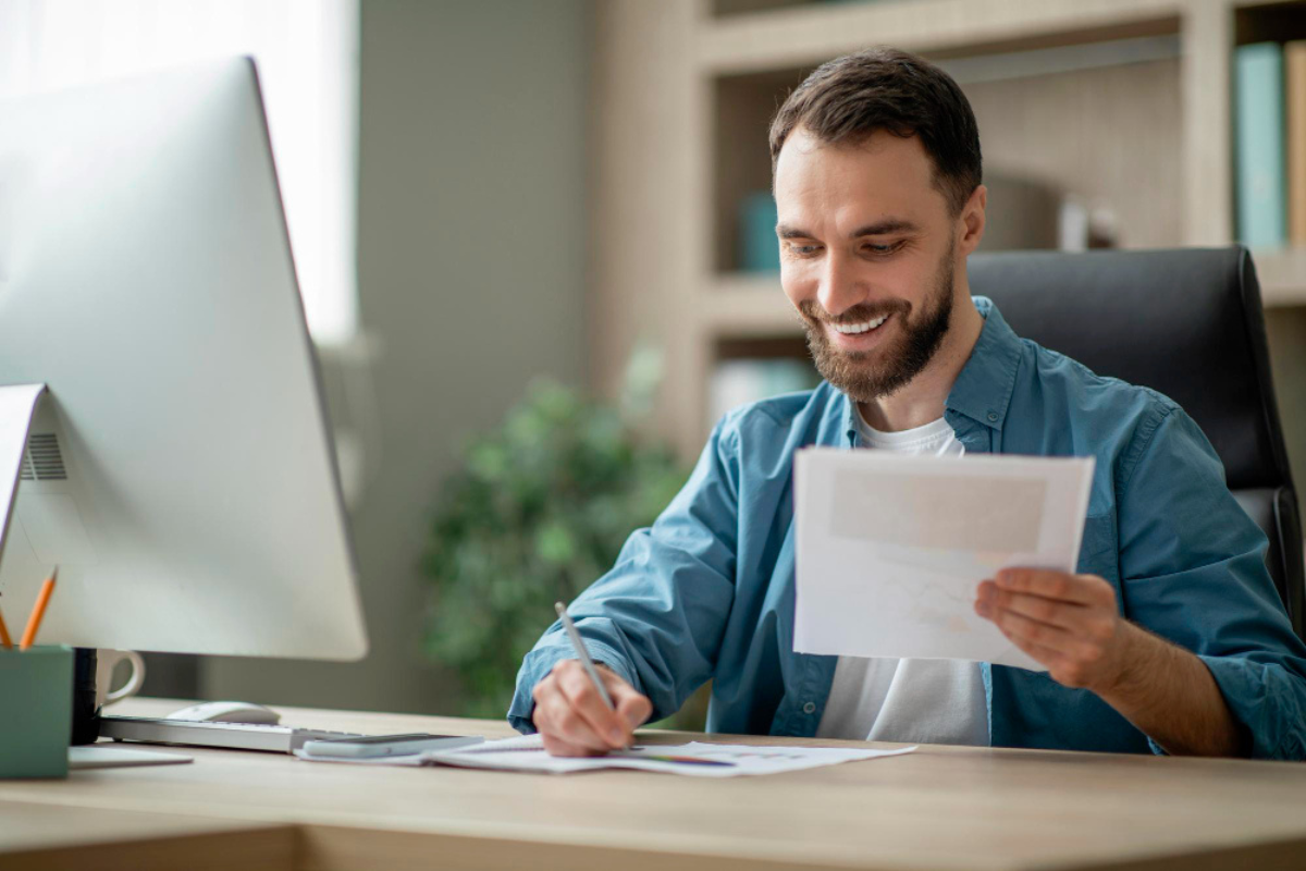 Homem sentado em frente ao computador segurando uma nota fiscal, com papéis sobre a mesa — imagem relacionada à emissão de nota fiscal para empresas prestadoras de serviço.