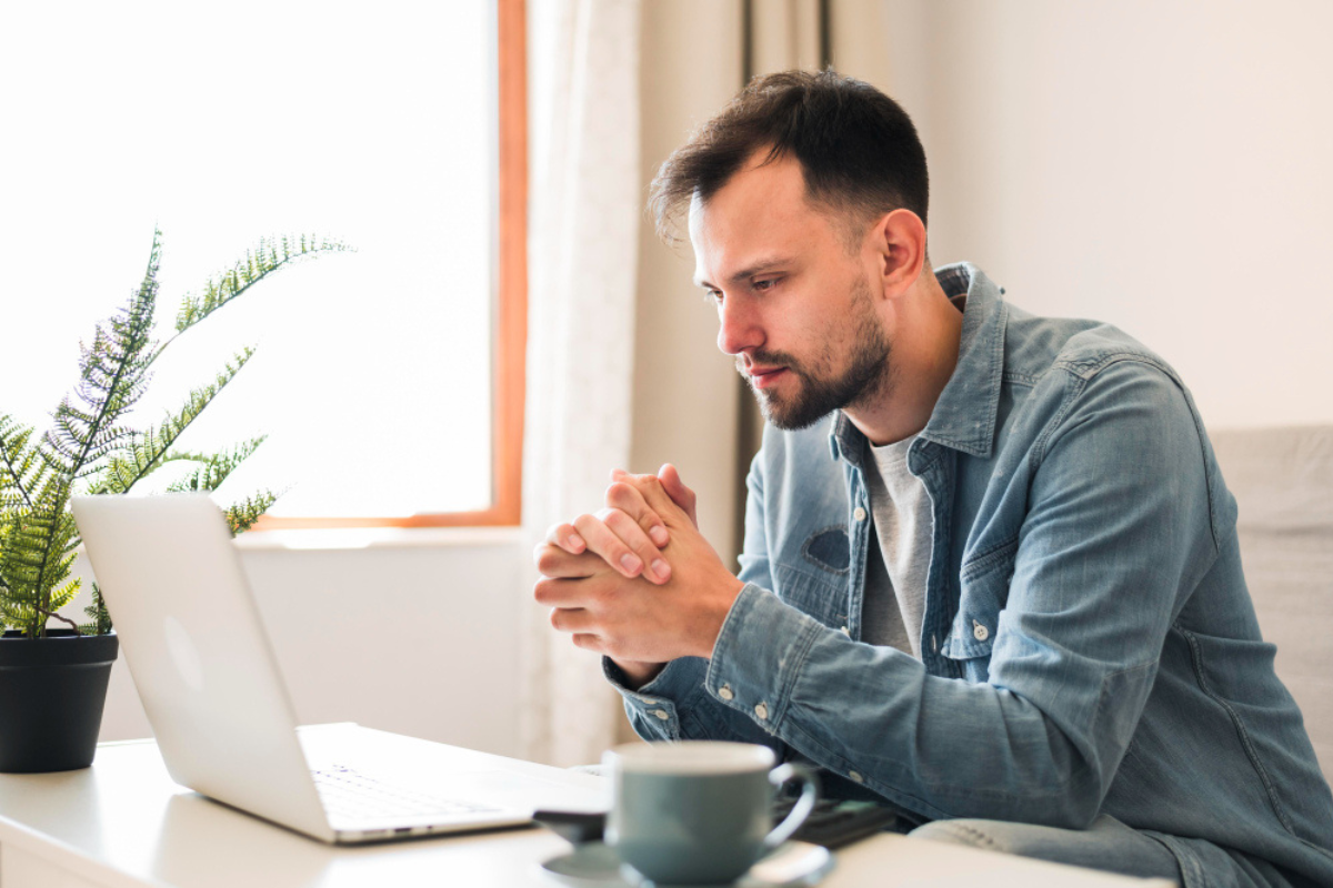 Homem sentado usando notebook sobre uma mesa de centro com uma xícara ao lado esquerdo e um vaso de plantas ao lado direito em ambiente de home office representando: CFOP 6107: venda de produção do estabelecimento, destinada a não contribuinte.