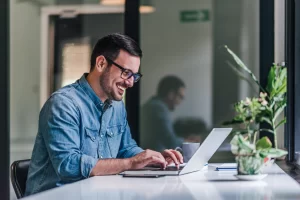 Homem sorridente em ambiente de escritório sentado usando um notebook sobre uma mesa com vaso de planta ao lado representando: CFOP 2202: devolução de venda de mercadoria adquirida ou recebida de terceiros.