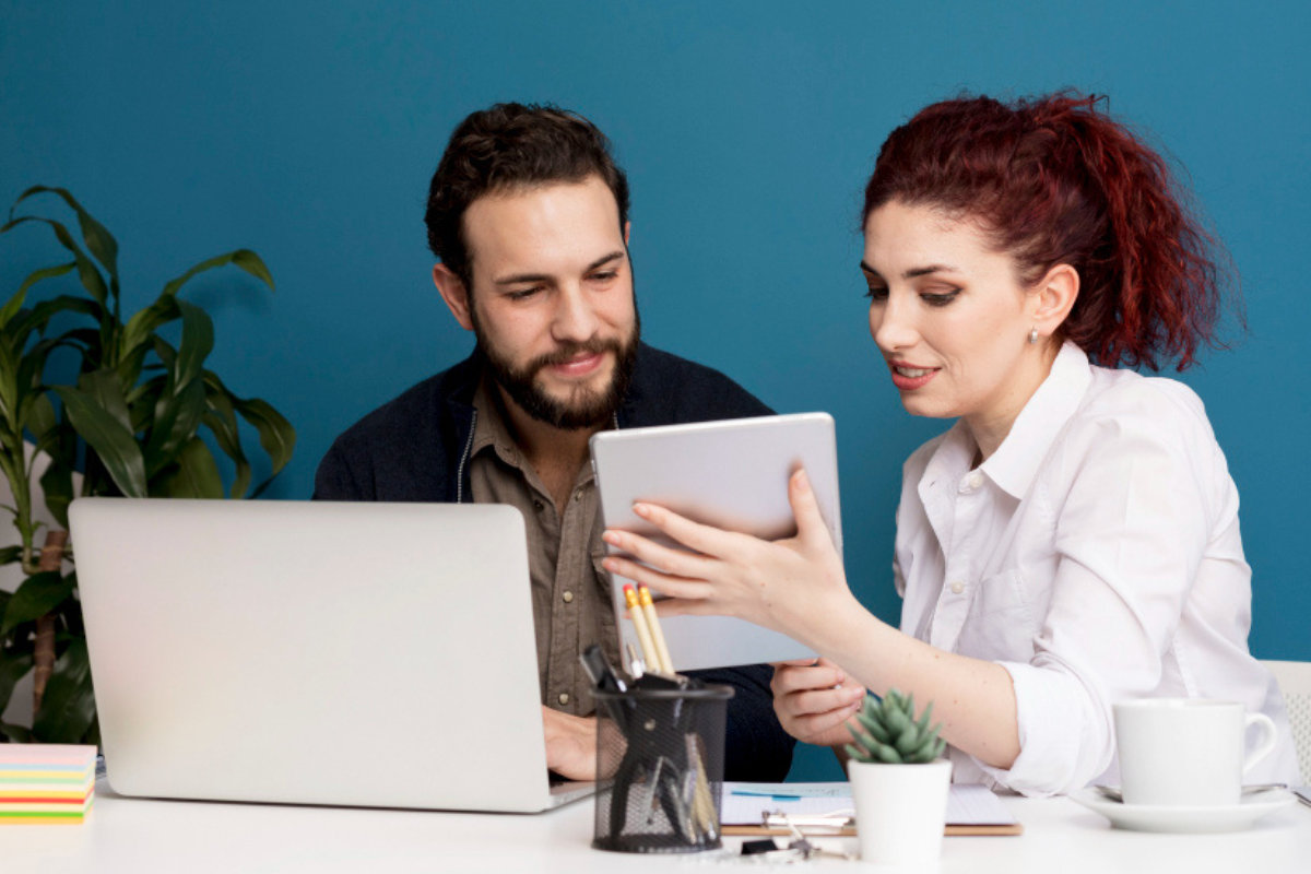 Homem e mulher em ambiente de escritório sentados em frente a um notebook que está sobre uma mesa próximo a uma xícara com pires, vaso de planta suculenta, bloco de anotações e um pote com canetas e lápis, conversando sobre algo que está na tela do tablet que se encontra nas mãos da mulher representando: Crédito fiscal de ICMS e PIS/COFINS: como funciona e o que pode ser abatido.