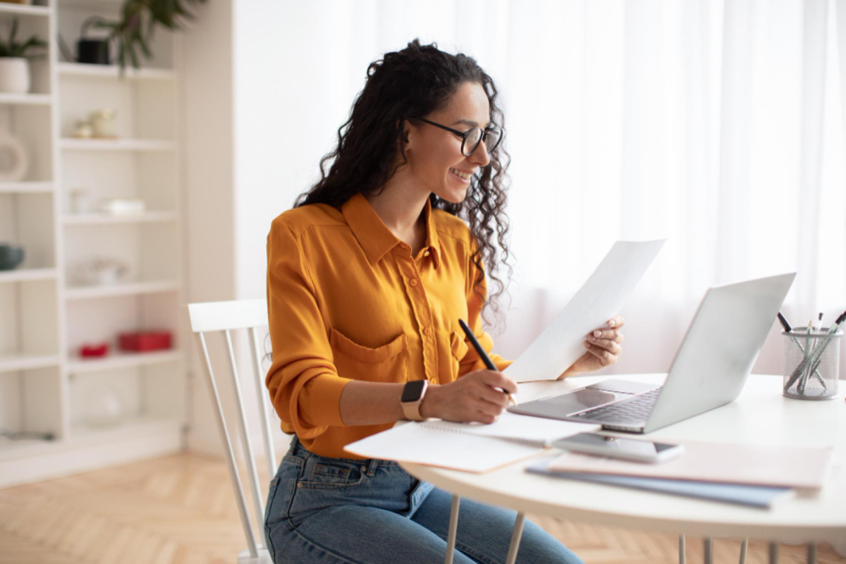 Mulher em ambiente de escritório home office sentada em frente um notebook sobra uma mesa com pote de canetas ao lado esquerdo, celular e dois blocos de anotações à direita, com uma caneta na mão direita fazendo anotações em um terceiro bloco e com um documento na mão esquerda representando: CFOP 1411: devolução de venda de mercadoria adquirida ou recebida de terceiros em operação com mercadoria sujeita ao regime de substituição tributária.