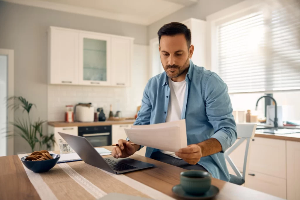 Home 15 Homem em ambiente de escritório home office sentando usando um notebook sobre uma mesa com uma xícara de café ao lado e um documento na mão esquerda representando: CFOP 1910: entrada de bonificação, doação ou brinde.