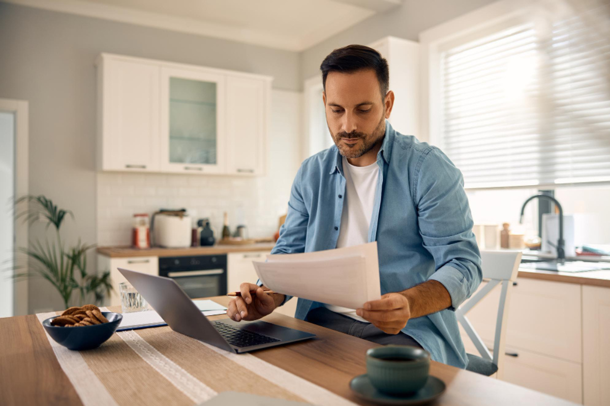 Homem em ambiente de escritório home office sentando usando um notebook sobre uma mesa com uma xícara de café ao lado e um documento na mão esquerda representando: CFOP 1910: entrada de bonificação, doação ou brinde.