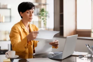 Mulher em ambiente de escritório sentada usando notebook sobre uma mesa com documentos em mãos representando: CFOP 5124: industrialização efetuada para outra empresa.