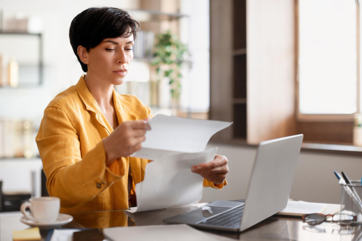 Mulher em ambiente de escritório sentada usando notebook sobre uma mesa com documentos em mãos representando: CFOP 5124: industrialização efetuada para outra empresa.