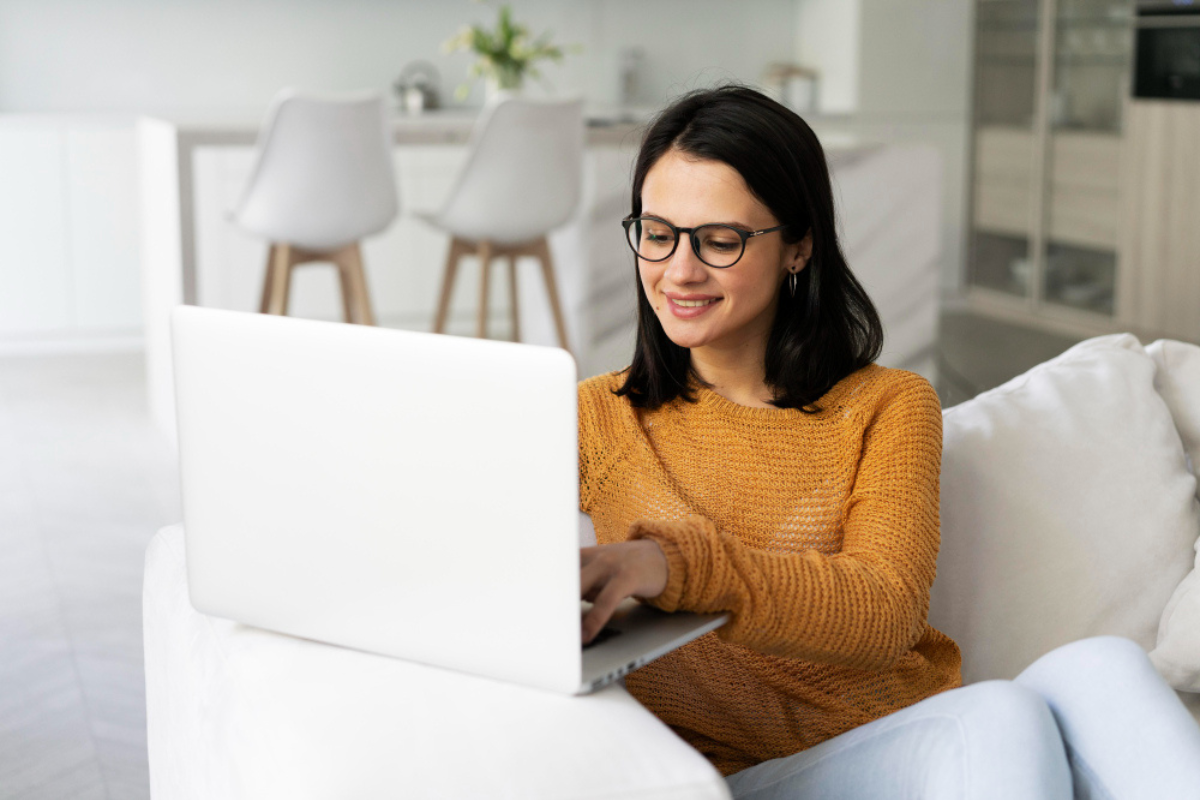Mulher senta em sofá usando notebook que está sobre o braço do sofá em um ambiente de sala de estar residencial com uma bancada e duas cadeiras ao fundo representando: Rejeição da nota fiscal: guia completo e principais tipos.