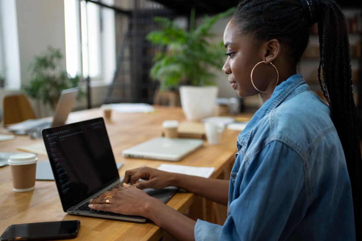 Mulher em ambiente de escritório sentada usando notebook sobe uma mesa com celular ao lado esquerdo e ao lado direito um espaço reservado com outro notebook para possível companheiro de equipe representando: Rejeição 938: não informada vBCSTRet, pST, vICMSSubstituto e vICMSSTRet.