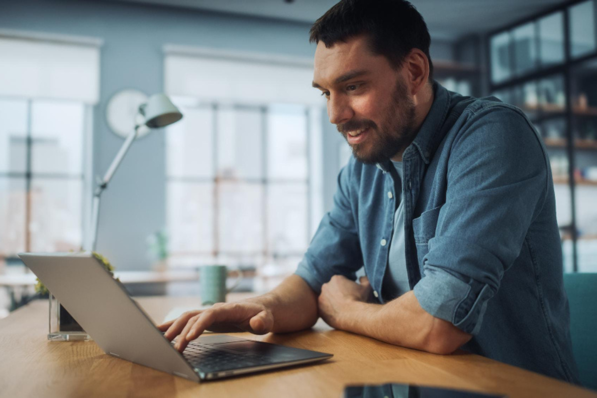 Homem em ambiente de escritório sentado usando notebook sobre uma mesa e ao lado um celular representando: Rejeição 539: duplicidade de NF-e, com diferença na Chave de Acesso.