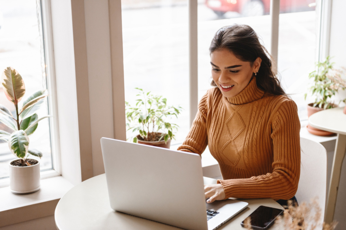 Mulher em ambiente de escritório em home office sentada usando notebook sobre uma mesa com três vasos de flores decorativas em torno representando: CFOP 1102: guia da compra para comercialização de produtos de origem no mesmo estado.