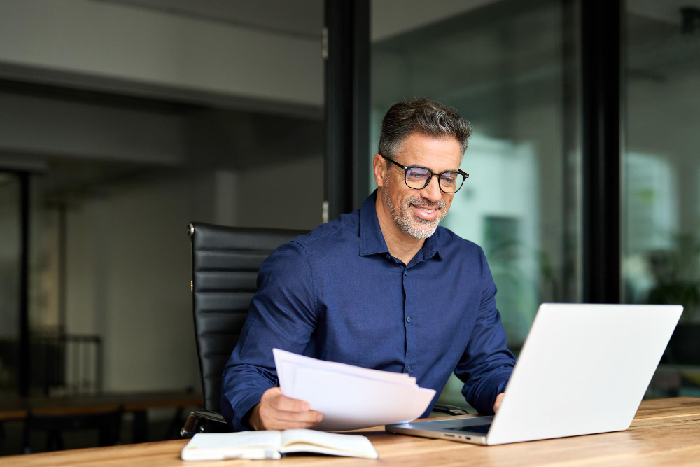 Homem em ambiente de escritório sentado usando notebook sobre uma mesa e lendo documentos em uma das mãos representando: Reforma Tributária: perguntas e respostas.