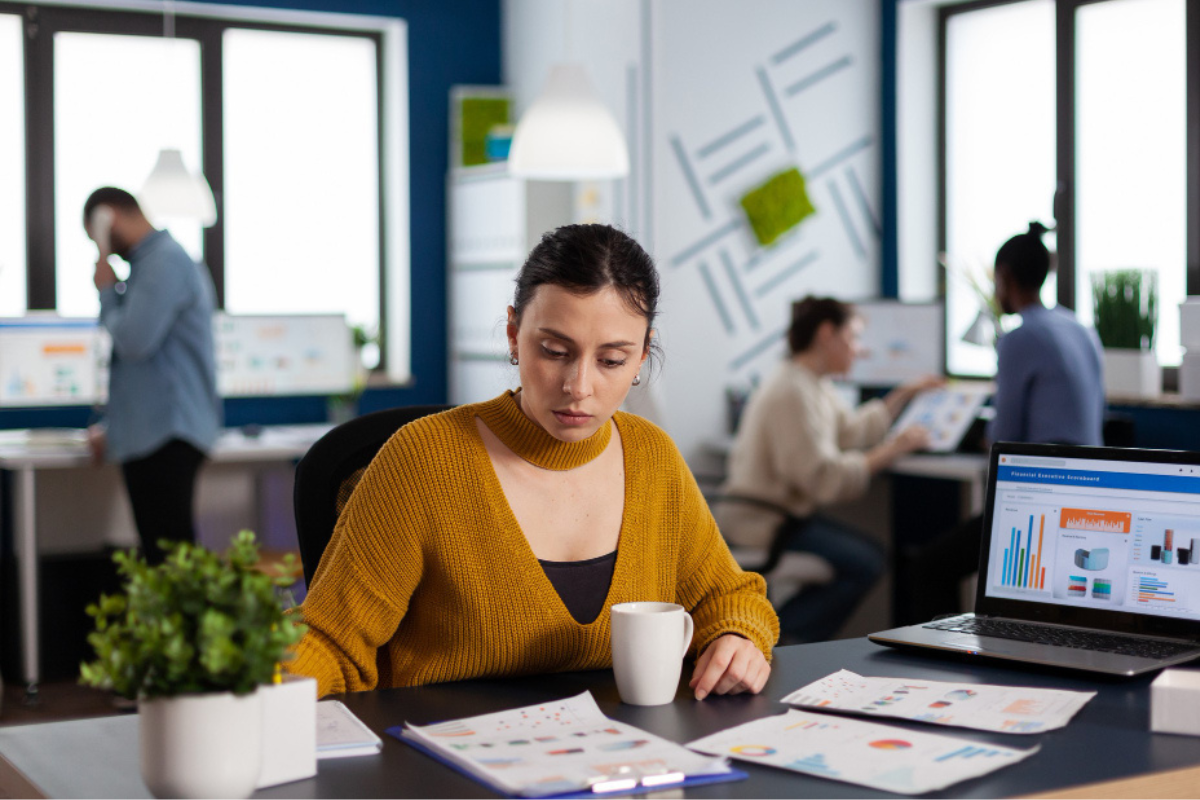 Mulher em ambiente de escritório sentada em frente a um computador analisando documentos sobre a mesa e ao fundo Três colegas de trabalho realizando a mesma tarefa representando: Tributação no destino: o que isso significa?