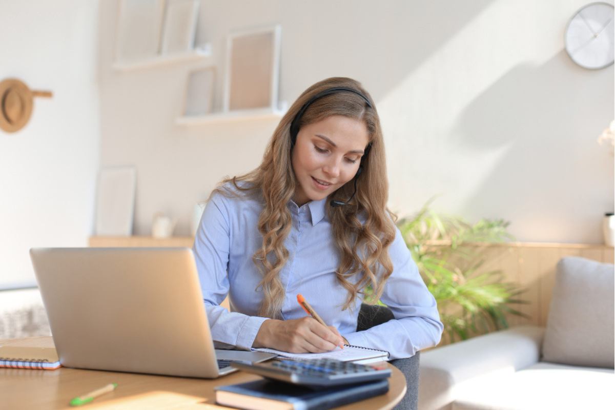 Mulher em ambiente de escritório sentada usando notebook sobre a mesa e fazendo anotações em uma agenda representando: A Reforma Tributária e o impacto na emissão de NF-e.