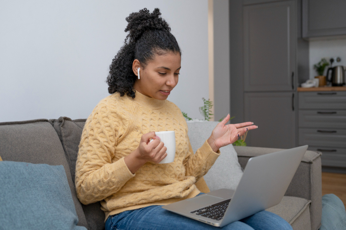 Mulher sentada em seu sofá de sua casa usando notebook no colo e com uma xícara em mãos representando: Glossário da Reforma Tributária: os principais termos explicados.
