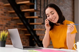 Mulher sorridente trabalhando em um notebook sobre uma mesa em ambiente de escritório e falando ao celular representando: Lucro presumido: o que é, alíquotas e como calcular.