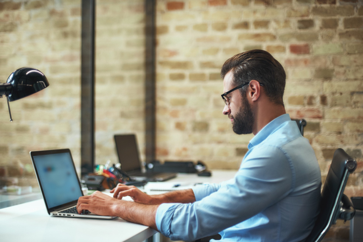 Homem sentado trabalhando em um notebook sobre uma mesa com mais pontos de trabalho ao lado em ambiente de escritório representando: 6 erros de gestão tributária que atrapalham sua empresa.