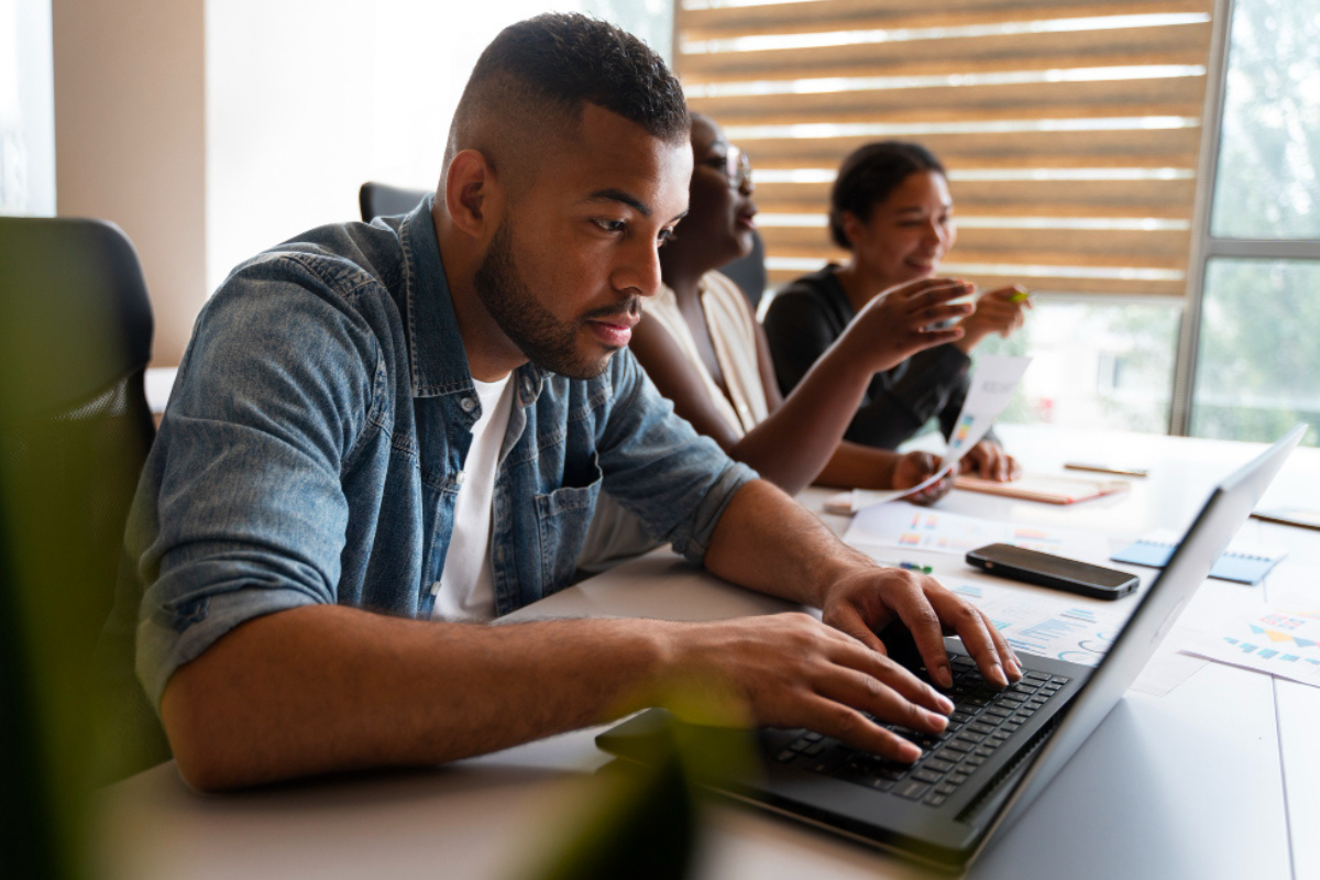 Homem sentado trabalhando em um notebook sobre uma mesa em ambiente de escritório com mais dois colegas de trabalho ao lado representando: Como preparar o emissor de notas fiscais para a Reforma Tributária em 2026.