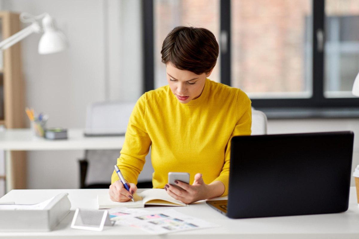 Mulher sentada em ambiente de escritório em frente a um notebook sobre uma mesa e olhando para um celular em uma mão e fazendo anotações em uma caderneta representando: O que é malha fina estadual e como evitar problemas com notas fiscais inconsistentes.