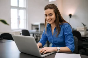 Mulher trabalhando em um notebook sobre uma mesa em ambiente de escritório representando: Como diferenciar o ICMS e o IBS na nota fiscal durante a transição da Reforma.