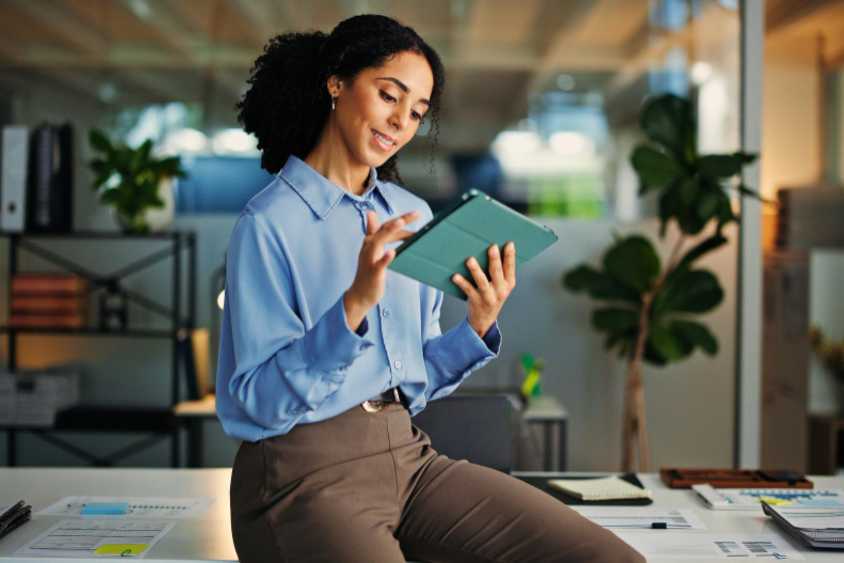 Mulher sentada na beirada da mesa usando um tablet em ambiente de escritório representando: CFOP 3556: compra de material para uso ou consumo.