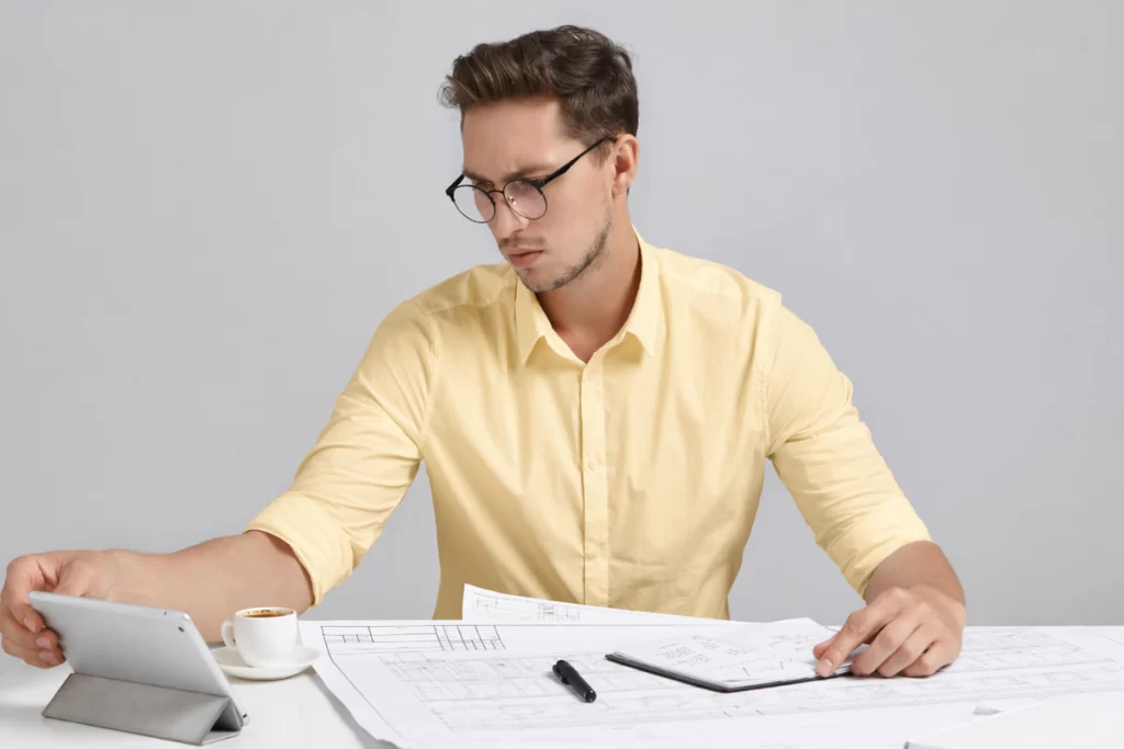 Homem sentado usando um tablet sobre uma mesa e fazendo anotações em agenda em ambiente de escritório representando: CFOP 3949: outra entrada de mercadoria ou prestação de serviço não especificado.