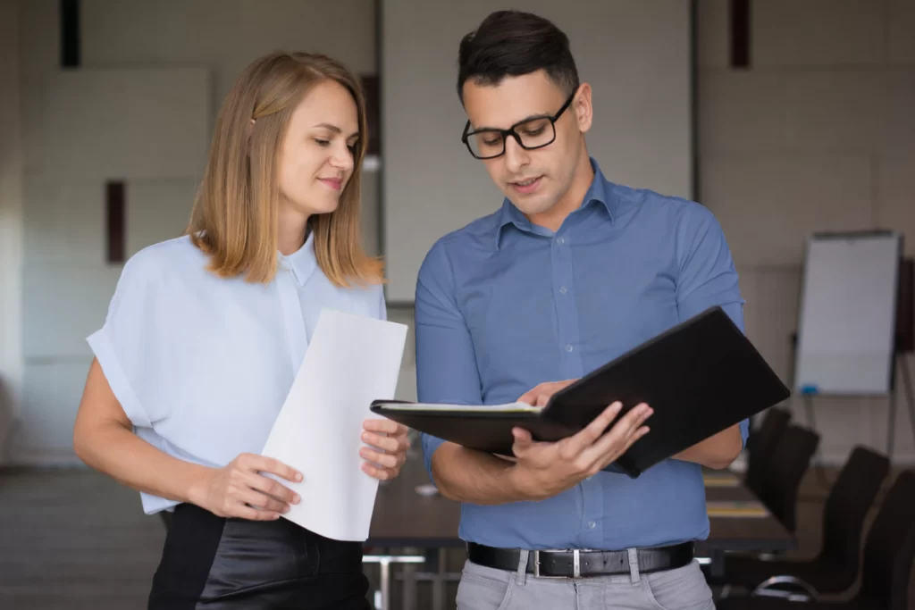 Em ambiente de escritório um homem com uma pasta arquivos e uma mulher com um documento em mãos conversam caminhando representando: CFOP 5214: Devolução de fixação de preço de produção do estabelecimento produtor.