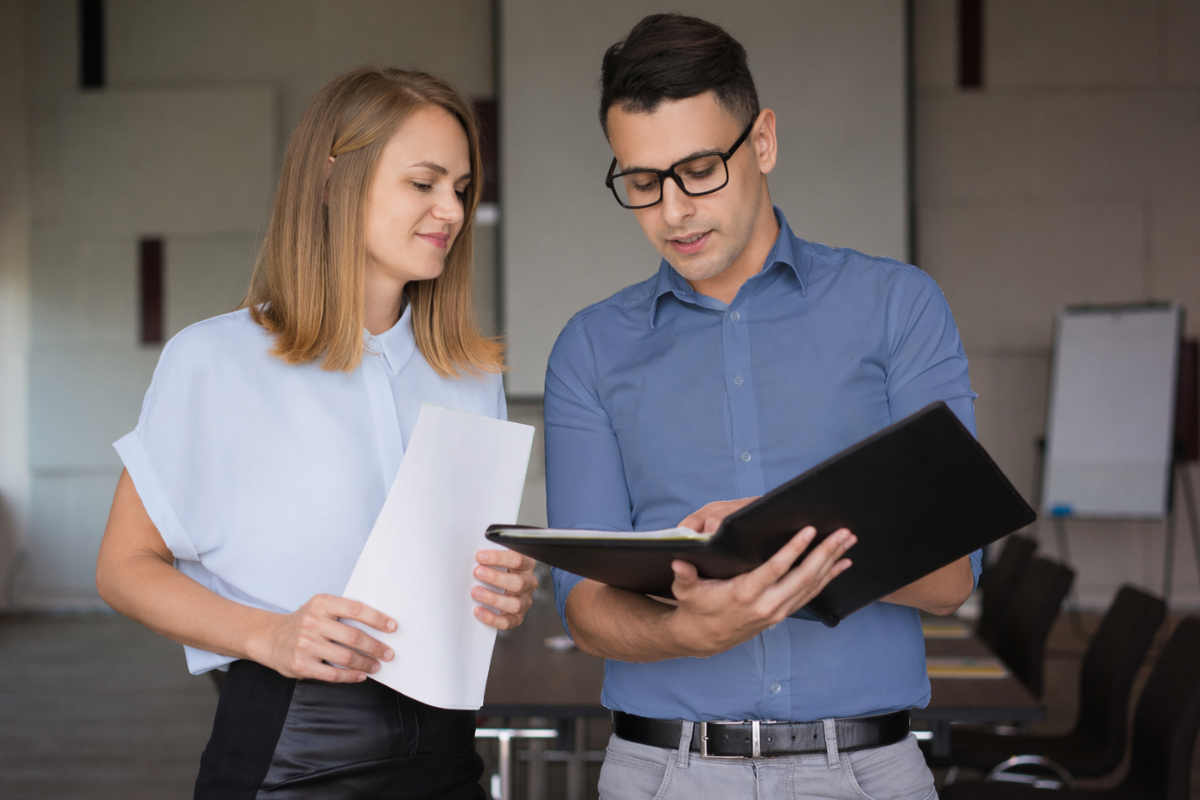Em ambiente de escritório um homem com uma pasta arquivos e uma mulher com um documento em mãos conversam caminhando representando: CFOP 5214: Devolução de fixação de preço de produção do estabelecimento produtor.