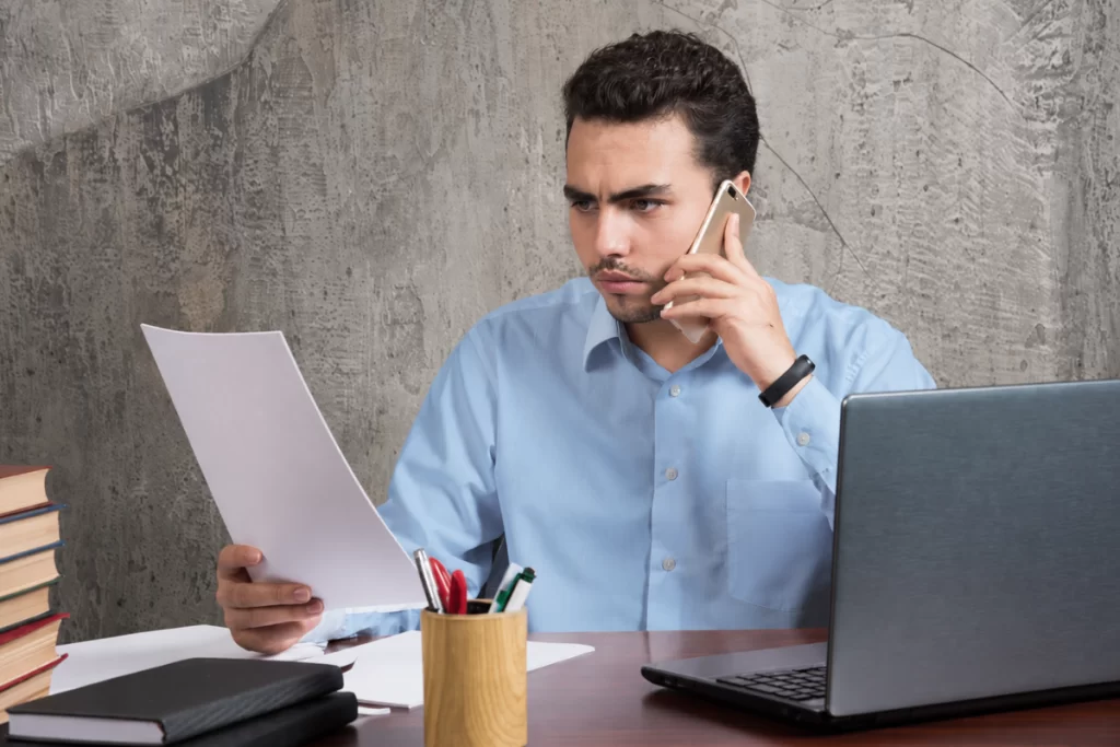 Profissional falando ao celular lendo um documento sentado em frente ao notebook sobre uma mesa em ambiente de escritório representando: O que acontece se sua empresa parar de emitir notas fiscais? Entenda os riscos.
