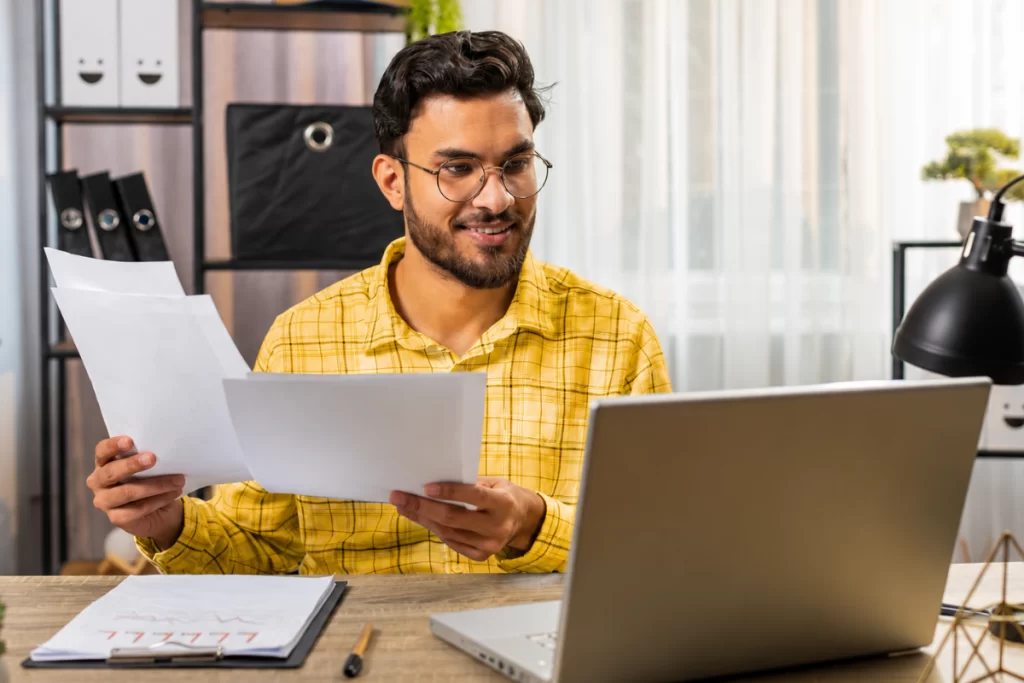 Homem analisando documentos em mãos e trabalhando em seu notebook em ambiente de escritório representando: Por que o estoque errado impacta imposto?