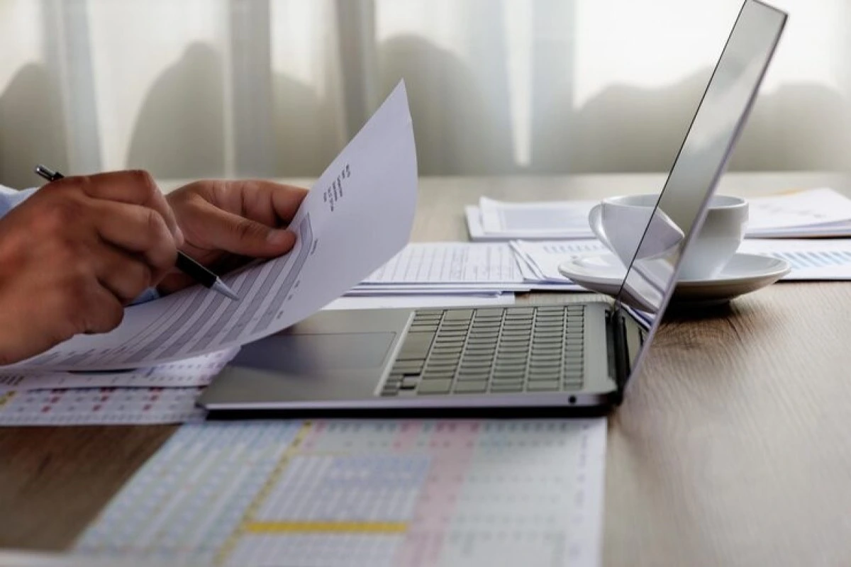 Homem de negócios trabalhando em seu notebook e fazendo anotações em documentos representando: Nota fiscal de honorários advocatícios: como emitir NFS-e corretamente
