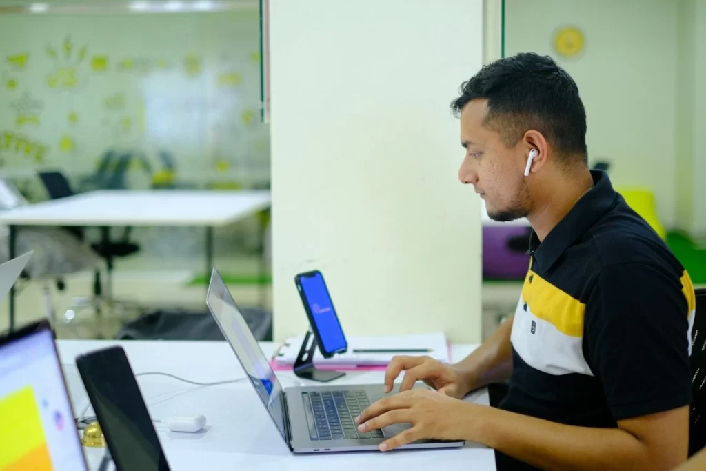 Profissional masculino em ambiente de escritório pesquisando em notebook e celular em uma mesa sobre Personal trainer pode ser MEI? CNAE certo, limite de faturamento e quando migrar para ME.