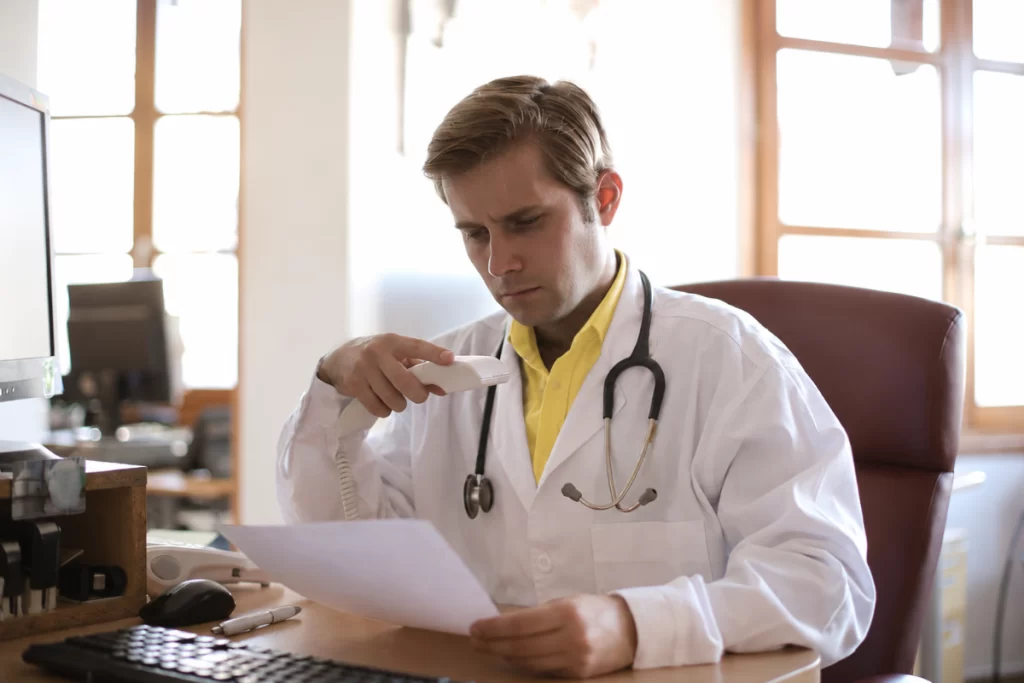 Médico na sua mesa de seu consultório lendo documento e com telefone fixo na mão representando: Médico pode ser MEI? Por que a maioria dos profissionais de saúde precisa abrir Simples Nacional