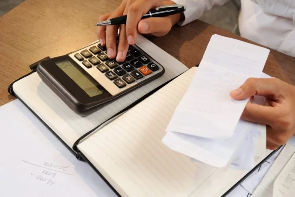 Homem usando calculadora, papel, caneta e fazendo anotações em agenda sobre uma mesa em ambiente de escritório representando: ISS para serviços de contabilidade: alíquotas, retenção e preenchimento da NFS-e.
